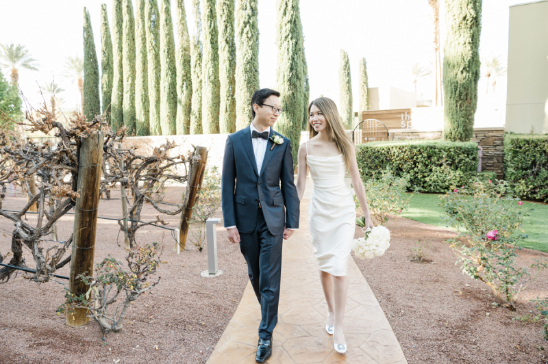 A groom and bride holding hands and walking up a sidewalk between rose bushes and grape vines.