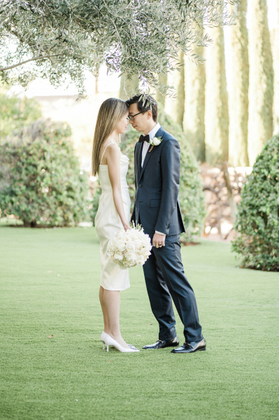 A bride and groom touch foreheads as they pose for wedding photos under a tree in a garden.