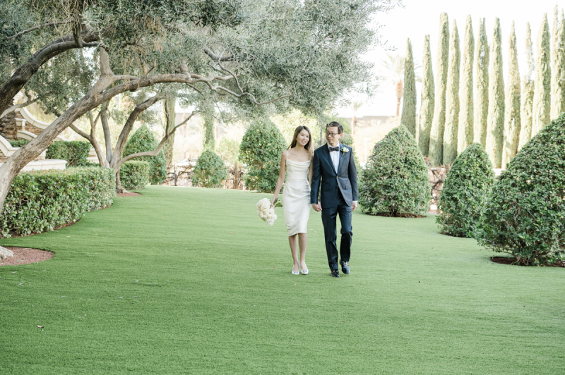A bride and groom holding hands and walking across a lawn in a garden.