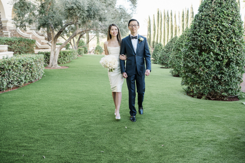 A groom escorts his bride as they walk towards the camera on a lawn in a garden.