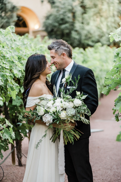Bride and groom holding a wedding bouquet, looking into each other\'s eyes and standing between rows of grape vines.