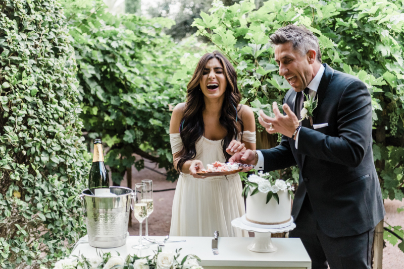 Bride laughing, groom licking fingers during wedding cake cutting celebration.