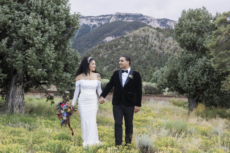 A bride and groom hold hands and turn their heads to look at each other as they stand in a meadow of wildflowers between pine trees with tree covered mountains in the background.