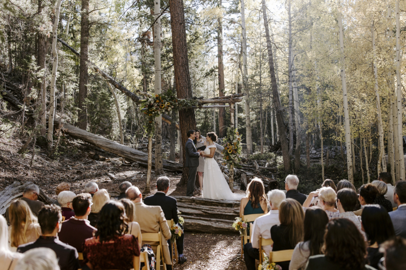 Wide shot of an outdoor wedding taking place in the mountains at Lee Canyon near Las Vegas. The wedding guests are seated on wooden folding chairs as they watch the bride and groom at a natural wooden arbor covered with wedding flowers.