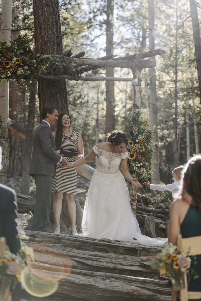 A bride reaches down to her ring bearer while her groom and officiant look on and laugh in a wooded mountain setting.