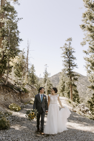 A bride and groom walk down a gravel road in the mountains with pine trees framing the road on both sides. He holds her floral bouquet while she holds the trail of her wedding dress.
