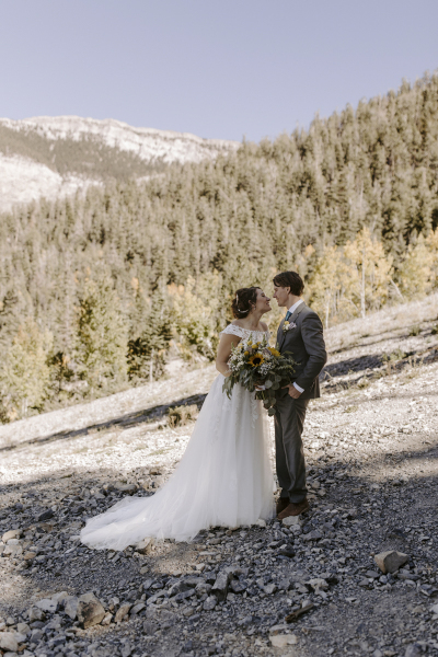 A bride and groom face each other lovingly as they are photographed on a rocky mountain slope with pine trees in the distance at Lee Canyon in Las Vegas.
