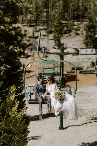 A bride and groom wearing sunglasses smile at each other and hold hands while riding the ski lift up the mountain at Lee Canyon ski area in Las Vegas on their wedding day.