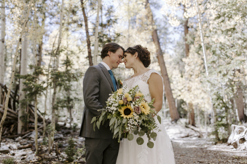 A bride and groom stand in a grove of aspen trees on their wedding day while the bride holds her bouquet of sunflowers, roses, eucalyptus and other flowers.