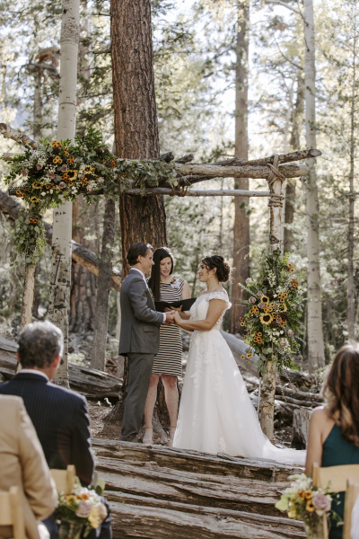 Wedding guests sit on wooden folding chairs as they watch a bride and groom hold hands and face each other and listen to their ceremony officiant standing underneath a wooden arbor decorated with flowers.