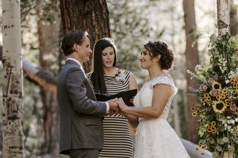 A bride and groom hold hands and face each other while listening to their officiant speak during their outdoor mountain wedding ceremony.