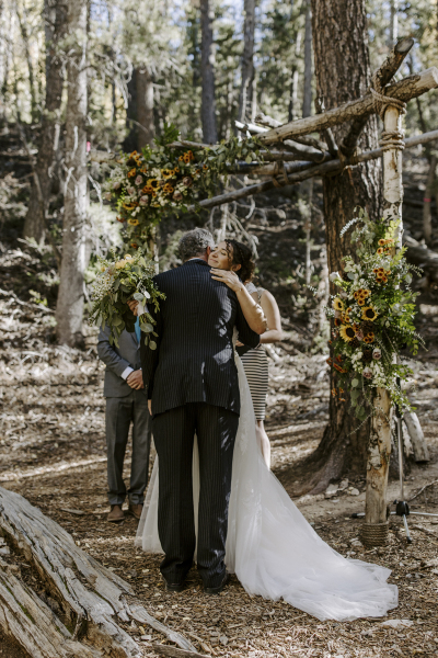 A man in a dark suit hugs a smiling bride while her groom waits for her under a wooden  arch covered in sunflowers .