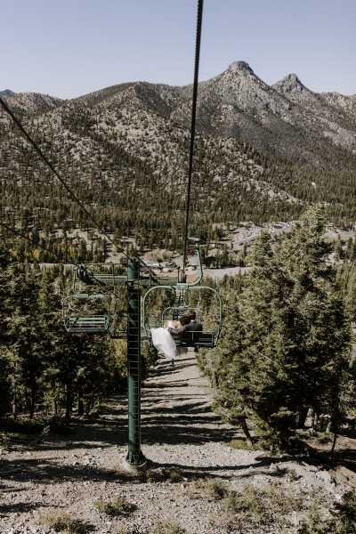 Wide shot of a bride and groom riding down the mountain on the ski lift at Lee Canyon ski area outside of Las Vegas. The bride rests her head on the groom's shoulder.