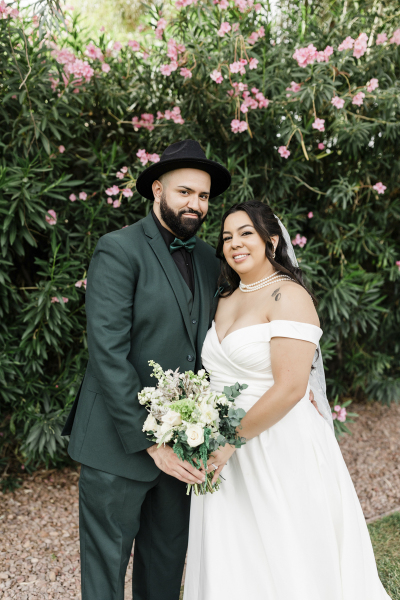 A bride and groom pose for the camera and smile as they stand in front of a row of pink Oleander bushes.