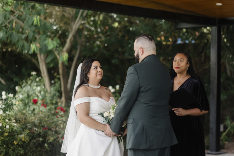 During a wedding ceremony, a bride looks at her groom who has his back to the camera. There is a rose garden in the background.