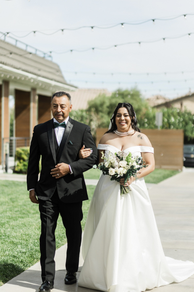 A bride is escorted by her father down a sidewalk in the backyard at the Lotus House in Las Vegas.