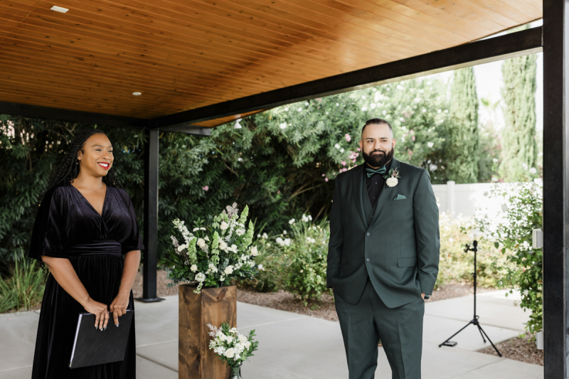 A groom and a wedding officiant look off camera and smile. They are standing under a garden pergola.