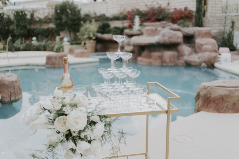 A gold-colored bar cart sits in front of a swimming pool and is decorated with white roses and a Champagne tower.