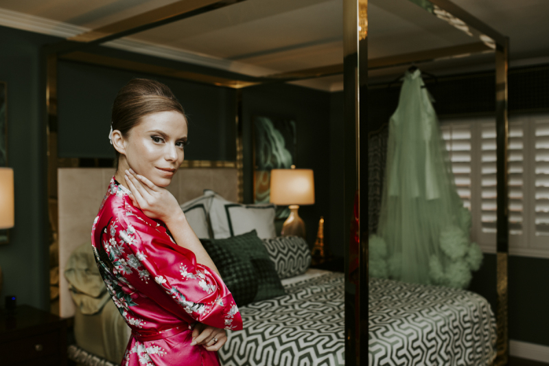 A woman looks over her shoulder as she stands in the bedroom of the Midcentury Manor in Las Vegas. Her light mint green wedding dress hangs at the far side of the four-poster bed.