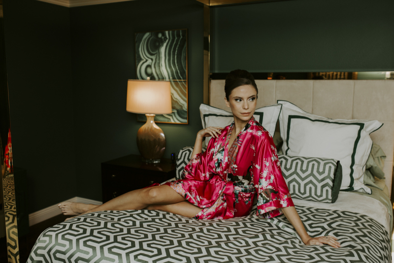 A woman in a pink silk floral-print bathrobe poses in a bedroom that is artfully decorated in midcentury modern style.