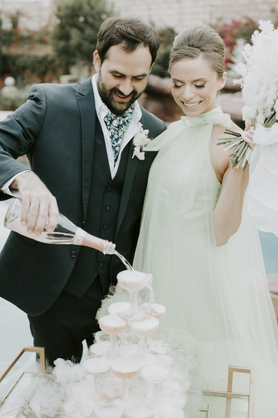 A groom pours pink Champagne into the top glass of a Champagne tower while his bride looks on and holds a bouquet of white flowers.