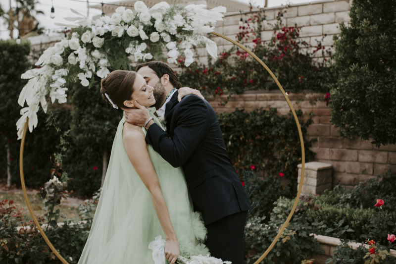 A groom kisses his wife's neck playfully on their wedding day in the garden of the Midcentury Manor in Las Vegas.