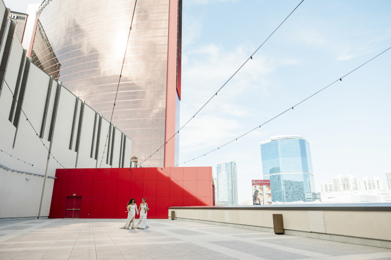 Two brides in elegant white gowns hold hands while strolling across a wide, modern terrace at Resorts World Las Vegas, with the resort’s striking red-and-glass tower and the blue skyline rising behind them.
