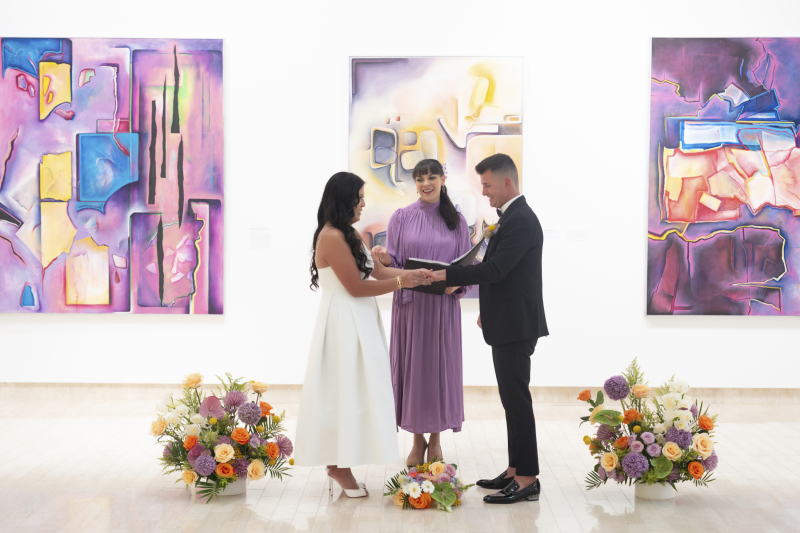 A bride places a wedding ring on her groom's finger during their wedding ceremony inside an art museum. as the officaint looks on.