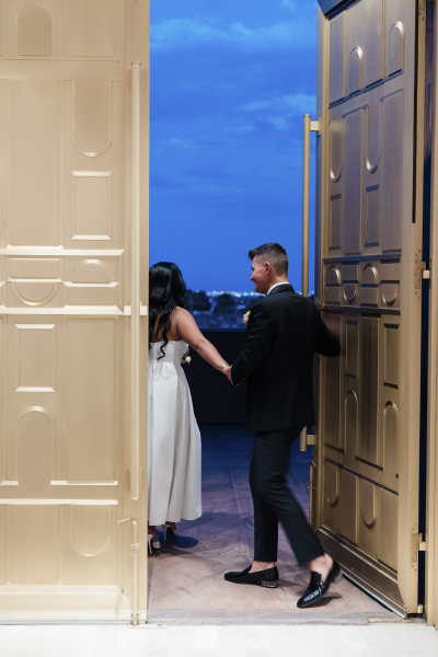 A groom follows his bride out a door to a patio with a view of the skyline.