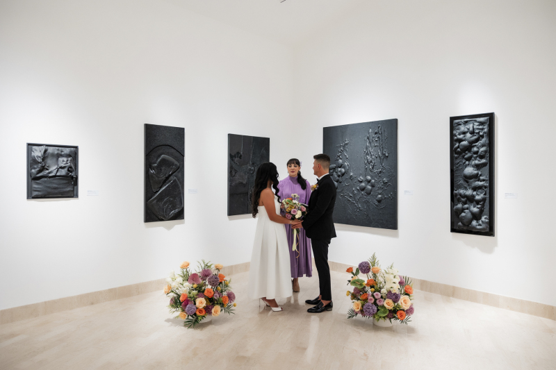 Standing between two floral bouquets a bride and groom get married in an art gallery as they hold hands and face their officiant.
