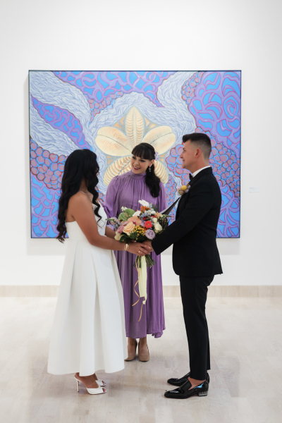 An officaint tilts her head to address the bride as a couple recites their vows in front of a large abstract flower painting.