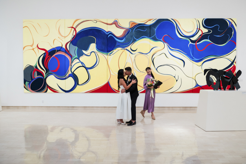 A newlywed couple share their first kiss as husband and wife  at the end of their wedding ceremony in an art gallery  with a giant yellow painting in the background.