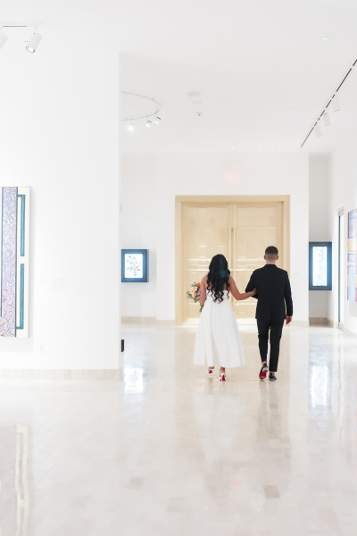 A bride and groom walk away from the camera as they move through the art museum towards a golden door.