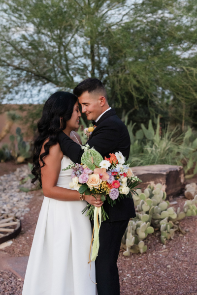In a cactus garden, a bride in a white dress and a groom in a black tuxedo close their eyes and bring their foreheads together. She holds a floral bouquet and he wraps his hand around her shoulder.