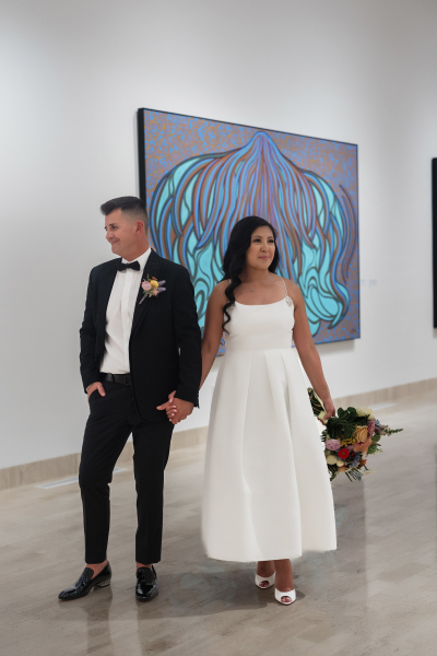 A groom and bride hold hands in an art gallery with a large teal painting behind them.