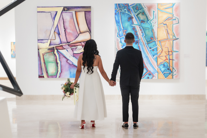 A bride and groom hold hands and stand facing away from the camera as they look at two large abstract paintings on the back wall of  the art museum where they got married.