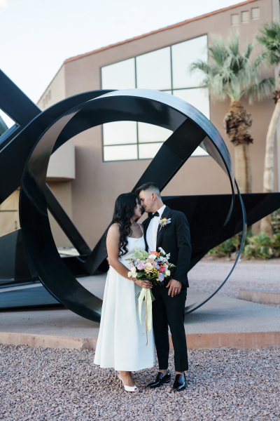 A bridge and groom stand in front of a large black metal sculpture .