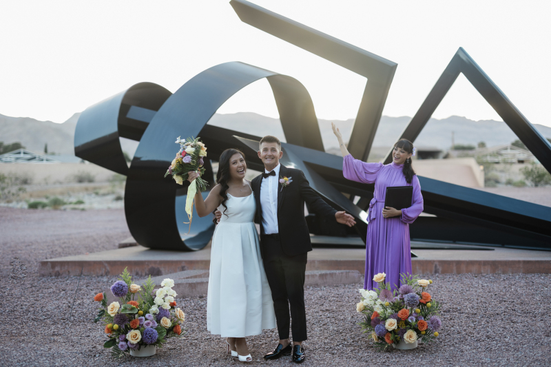 A bride, groom and officiant raisie their hands in celebration as the couple gets married in front of a large black abstract outdoor sculpture