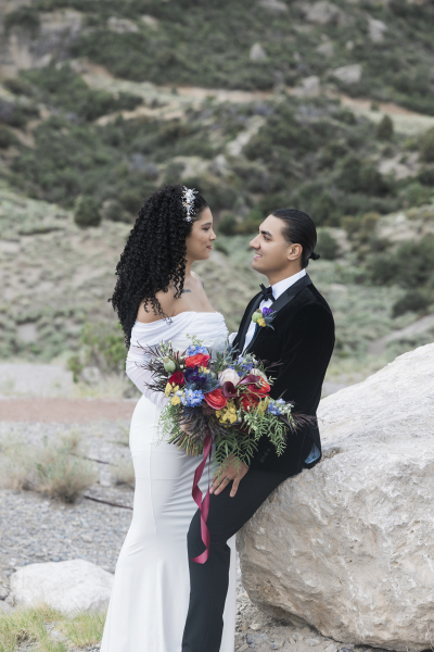 A groom sits on a boulder as his bride stands between his legs. The couple looks lovingly at each other and smiles.
