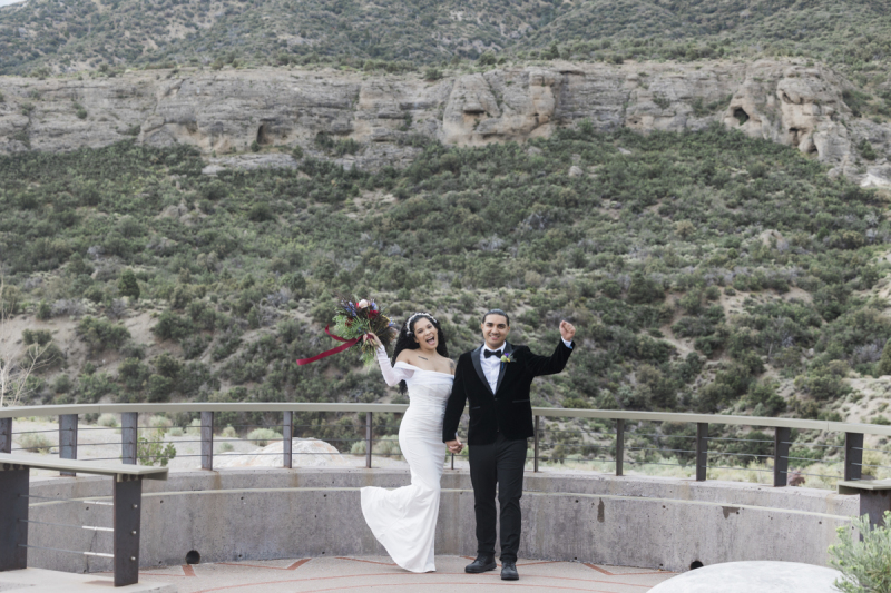 A bride and groom raise their arms in celebration of their marriage in the Spring Mountains outside of Las Vegas as they stand in front of a hillside dotted with a large rock outcropping.