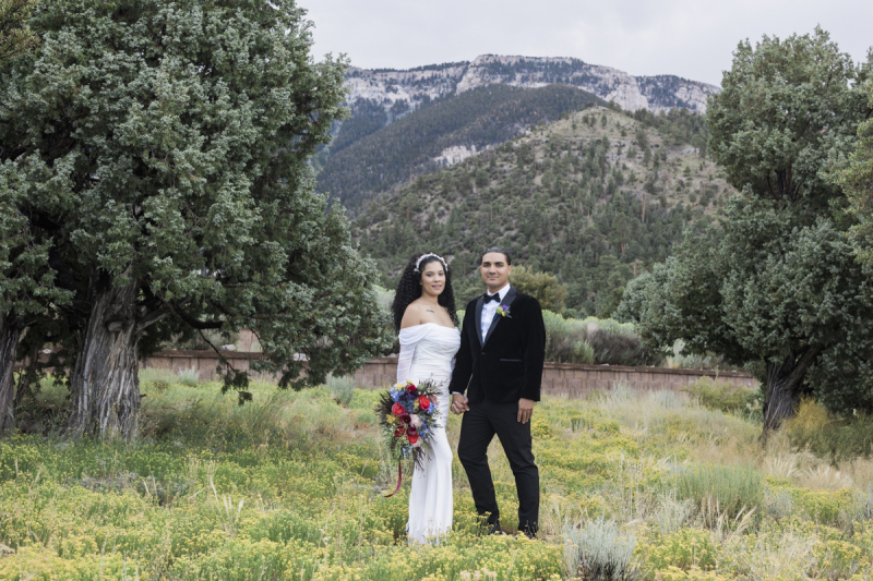 A bride and groom take a relaxed and casual stance as they pose in a meadow of wildflowers between pine trees with tree covered mountains in the background.