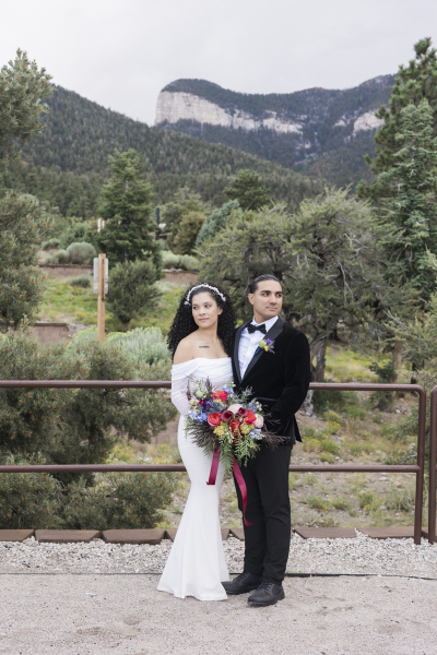 A medium shot of a bride and groom holding a bouquet of flowers between them as they turn their heads in opposite directions to look at the surroundings of pine trees and mountains.