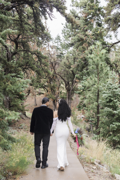 A groom and bride walk hand in hand away from the camera down a paved path through the pine trees, blooming bushes and tufted grasses.