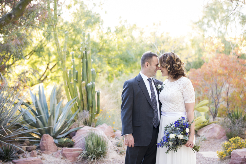 A middle-aged groom and bride press their noses together on their wedding day at Springs Preserve in Las Vegas. They are standing in a rocky desert garden filled with cacti.