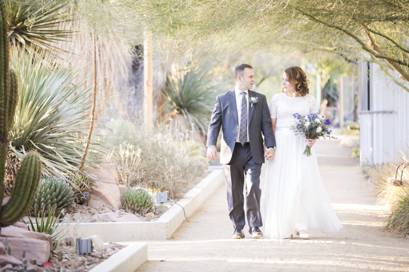 A groom and bride hold hands while strolling along a path in a desert garden. They are looking at each other as sunlight streams in from their right.