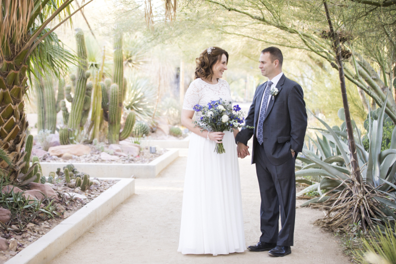 A bride and groom stand facing each other and holding hands. They are underneath a palm tree planted amongst some large cacti and agave plants growing at the Springs Preserve in Las Vegas.