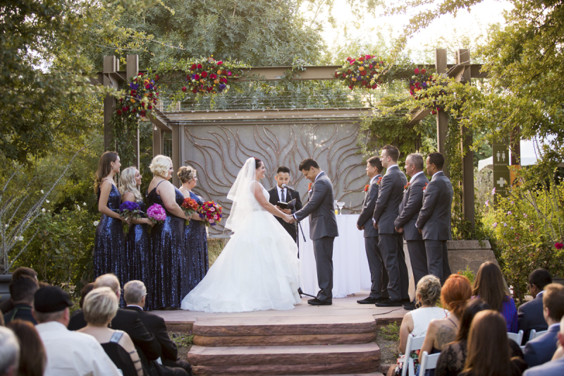 A desert garden wedding is being held underneath an arbor decorated with flowers. There is a sunburst sculpture behind the arbor. 