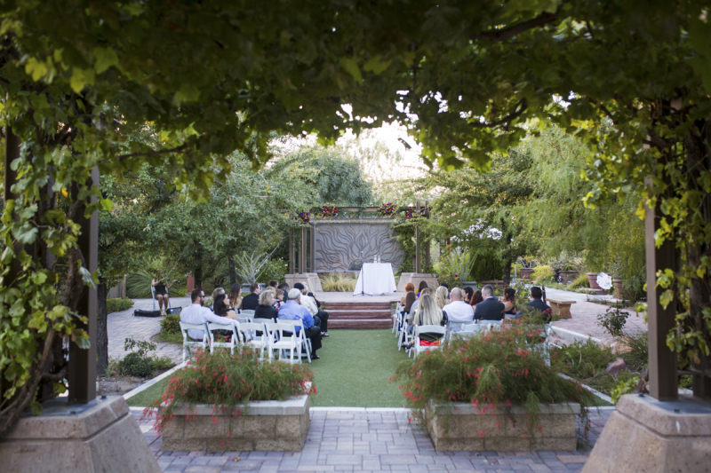 Wedding guests sit in white wooden folding chairs in a garden waiting for the ceremony to begin. The garden is surrounded by trees, bushes, and grape vines and framed by stone pavers.