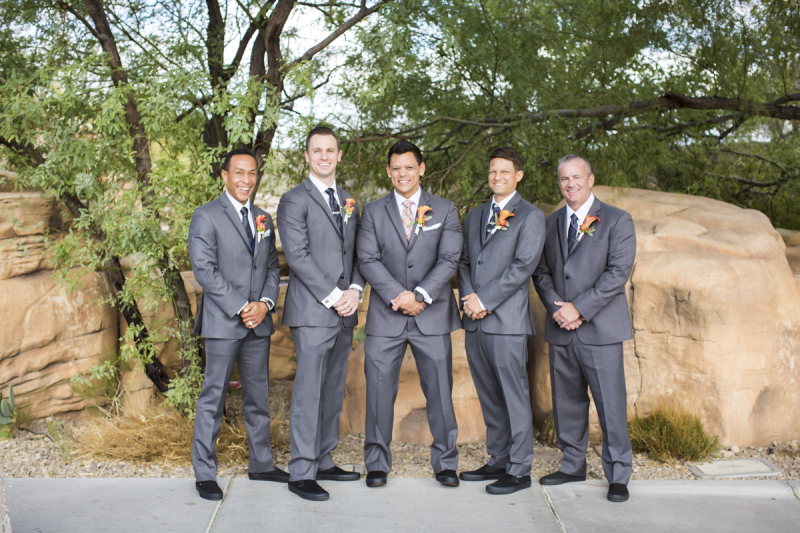 A groom and his groomsmen pose in front of a rock outcropping at Springs Preserve in Las Vegas. They are wearing grey suits and matching black shoes and each clasp their hands in front of them at their waist.