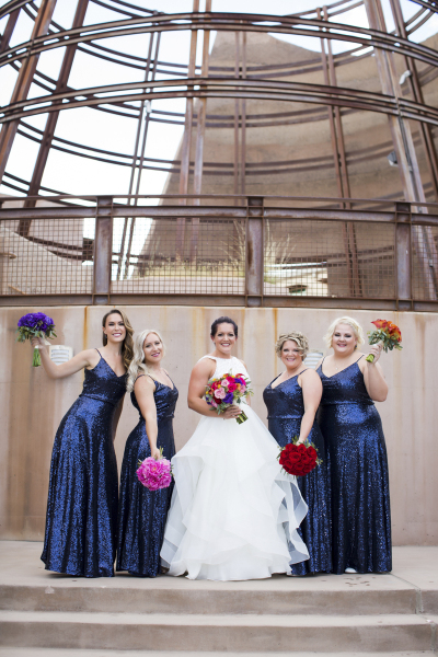 A bride in a white wedding dress poses with her bridesmaids. The bridesmaids are wearing sparkly blue dresses and each have a different colored floral bouquet. The bride has flowers from each of the other four bouquets in her bouquet. They are standing in front of a concrete and steel beam sculpture. 